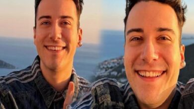 Two men smiling for a selfie on a coastal overlook at sunset, with the sea and cliffs in the background, wearing denim jackets.