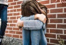 Girl sits against a brick wall with knees drawn up, arms wrapped around them, looking distressed or sad.