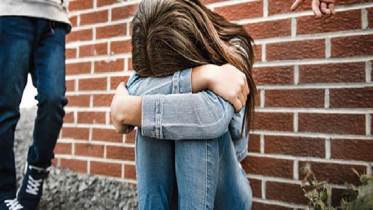 Girl sits against a brick wall with knees drawn up, arms wrapped around them, looking distressed or sad.