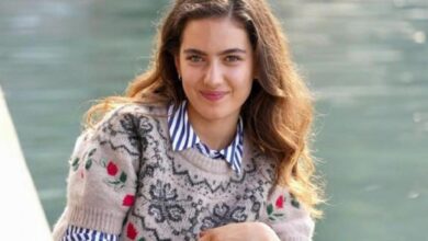 Smiling young woman with wavy brown hair wearing a floral-pattern knit sweater over a striped shirt by a waterway.
