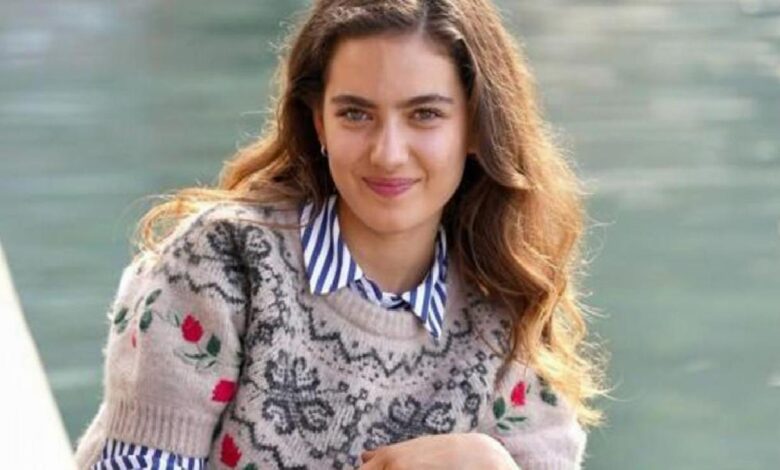 Smiling young woman with wavy brown hair wearing a floral-pattern knit sweater over a striped shirt by a waterway.