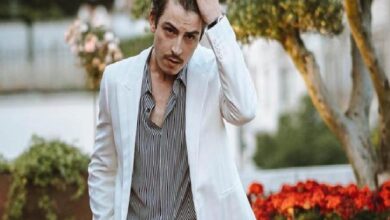 Man in a white blazer and striped shirt, brushing his hair, standing in a garden with red flowers in the background.