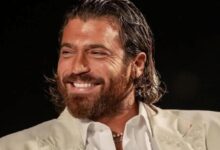Man with dark hair and beard, wearing a white suit, smiling against a black background.
