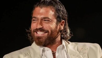 Man with dark hair and beard, wearing a white suit, smiling against a black background.