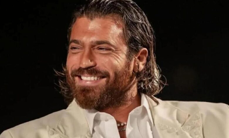 Man with dark hair and beard, wearing a white suit, smiling against a black background.