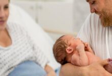 Newborn baby cradled in father's arms as mother lies in a hospital bed nearby in a maternity room.
