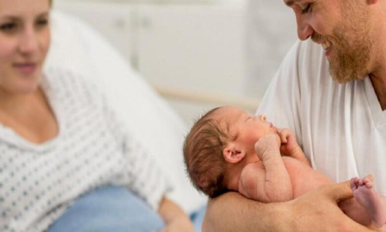 Newborn baby cradled in father's arms as mother lies in a hospital bed nearby in a maternity room.