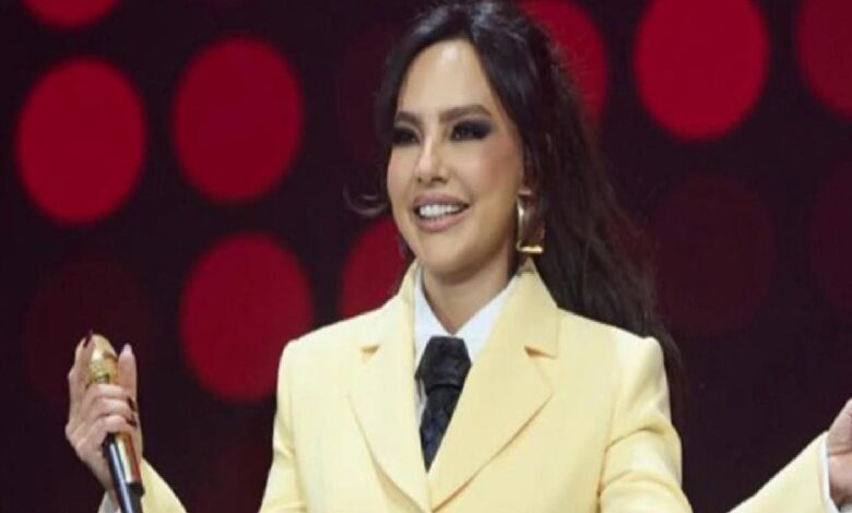 Woman on a stage wearing a pale yellow suit and black tie, holding a microphone and smiling.