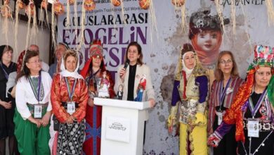 Group of people in traditional costumes on a stage, with a woman at a podium speaking into a microphone, banner behind them.