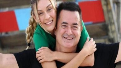 Smiling man being hugged from behind by a woman in green, outdoors, both looking at the camera in a joyful pose.