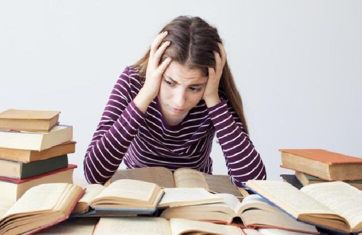 Young woman sits at a desk surrounded by open books, holding her head in frustration while studying.