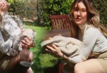 Two women outdoors holding babies wrapped in blankets in a sunny garden setting.