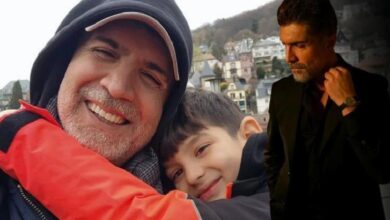 Smiling older man in a red jacket and cap hugs a boy; a man in a black suit stands nearby, against a hillside town backdrop.