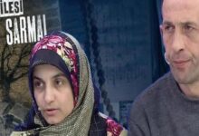 Two people sit side by side on a TV set; a woman in a colorful headscarf speaks while a man in a gray sweater listens in the background.