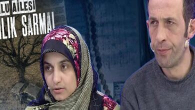 Two people sit side by side on a TV set; a woman in a colorful headscarf speaks while a man in a gray sweater listens in the background.