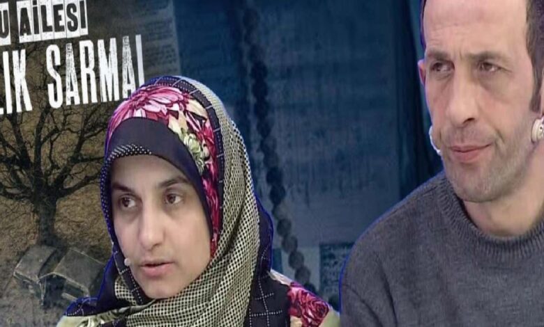 Two people sit side by side on a TV set; a woman in a colorful headscarf speaks while a man in a gray sweater listens in the background.