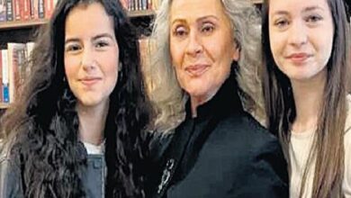 Three women pose for a photo in a library: an older woman in a black jacket at center, flanked by two younger women with long dark hair.
