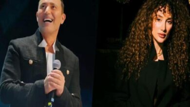 Two performers on stage: a man in a dark jacket holding a microphone on the left and smiling, with a woman with long curly hair in a dark outfit on the right.