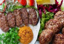 Plate with grilled kofta kebabs on flatbread, fresh herbs, and orange bulgur; crispy fried meat croquettes on greens nearby.