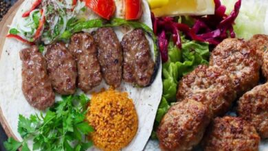 Plate with grilled kofta kebabs on flatbread, fresh herbs, and orange bulgur; crispy fried meat croquettes on greens nearby.