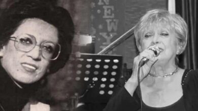 Two women on a stage: left woman wears glasses and a hat, right woman sings into a microphone.