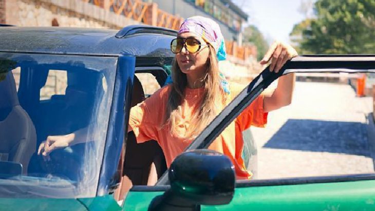 Woman wearing a headscarf and sunglasses leaning out of an open car window on a sunny day.