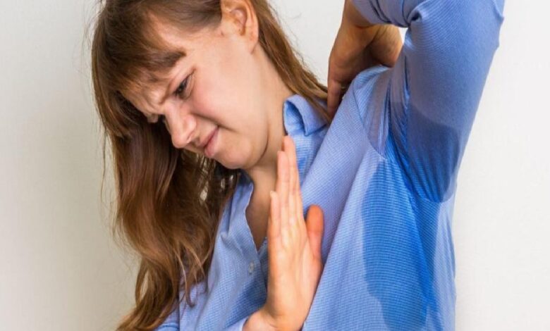 Woman in a blue shirt performs a shoulder stretch, raising an arm behind her head with hands pressed together in front of her chest.