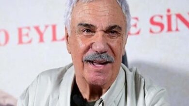 Close-up of an older man with gray hair and mustache speaking, wearing a light shirt, with red text in the background.