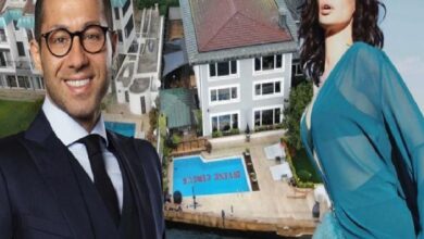 Smiling man in a dark suit and glasses and a woman in a blue dress posing in front of a luxury property with a pool behind them.