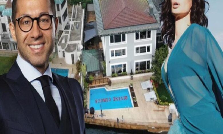 Smiling man in a dark suit and glasses and a woman in a blue dress posing in front of a luxury property with a pool behind them.