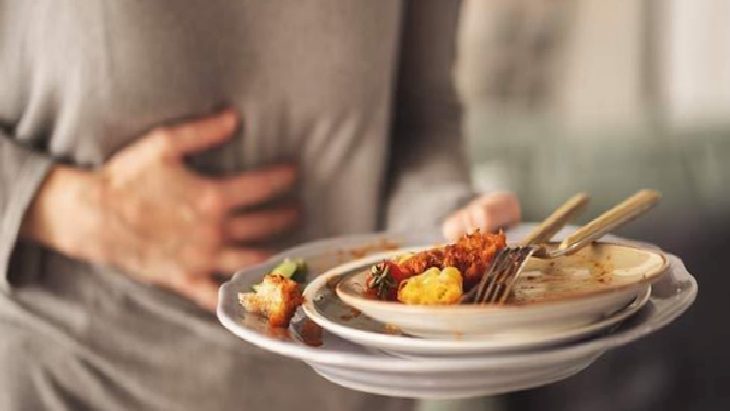 Person in a gray top holds a plate with partly eaten food while clutching their abdomen in apparent stomach pain.