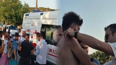 Shirtless man being attended to by another person near an ambulance as onlookers stand by outdoors; faces blurred for privacy.