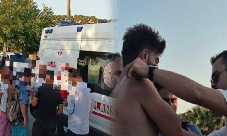 Shirtless man being attended to by another person near an ambulance as onlookers stand by outdoors; faces blurred for privacy.