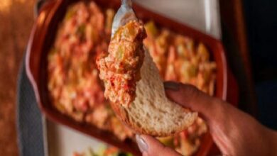 Close-up of a hand holding a crusty bread slice dipped into a creamy tomato-and-vegetable spread in a baking dish behind.