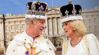 Two older adults in royal costumes and crown-like hats chat in front of a grand palace facade.