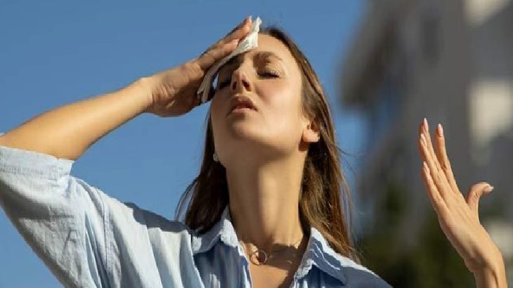 Woman outdoors in a light blue shirt wipes sweat from her forehead with a tissue under a bright blue sky.