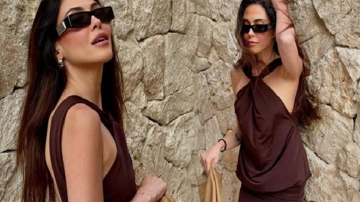Two women in brown outfits and sunglasses pose against a rough stone wall, one holding a beige bag.