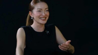 Smiling woman in a black sleeveless top gestures with her right hand; lavalier microphone clipped to her chest against a dark background.