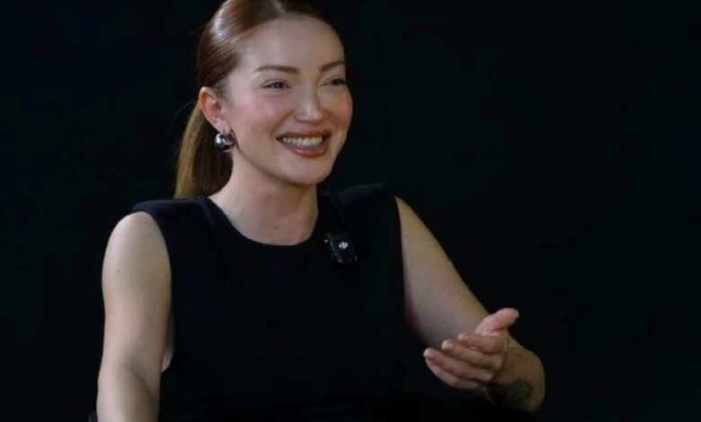 Smiling woman in a black sleeveless top gestures with her right hand; lavalier microphone clipped to her chest against a dark background.