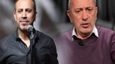 Two men on a stage, one at a microphone wearing a black shirt, the other in a purple sweater looking serious.