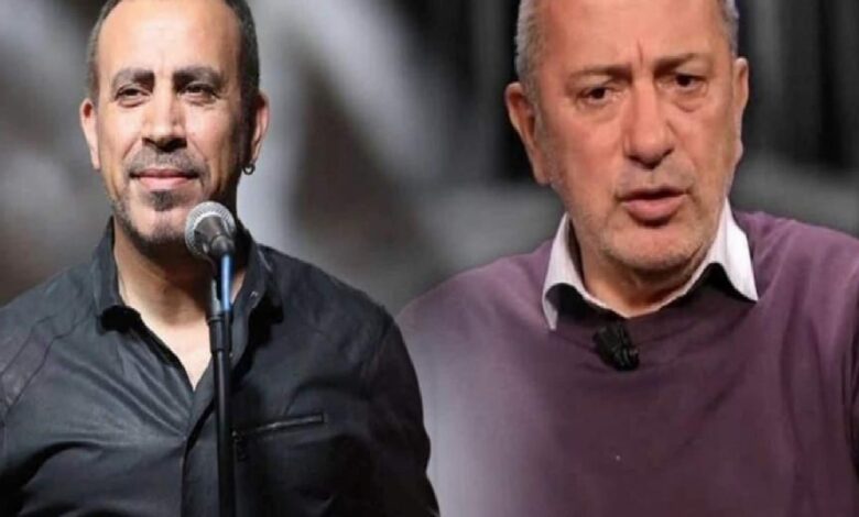 Two men on a stage, one at a microphone wearing a black shirt, the other in a purple sweater looking serious.