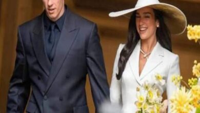 A smiling couple walking together; woman in a white suit and wide-brim hat holding flowers, man in a dark suit beside her.