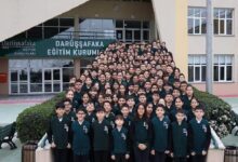 Large group of students in dark green uniforms posing on the stairway outside a school entrance with a banner reading 'Darüşşafaka Eğitim Kurumu'.