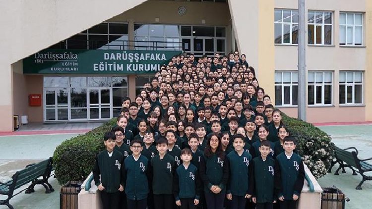 Large group of students in dark green uniforms posing on the stairway outside a school entrance with a banner reading 'Darüşşafaka Eğitim Kurumu'.