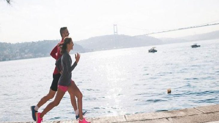 Two runners, a man in red and a woman in gray with pink shorts, jog along a waterfront with a bridge in the distance.