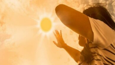 Person outdoors on a sunny day, raising an arm to shield their eyes from bright sunlight.