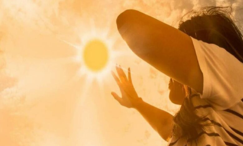Person outdoors on a sunny day, raising an arm to shield their eyes from bright sunlight.