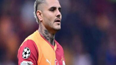 Soccer player with neck tattoos in a red-and-orange jersey on the field during a match, Champions League patch visible on sleeve.