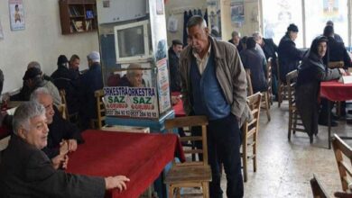 Busy cafe with red-tablecloths; elderly patrons chat while a man stands beside a pillar with an orchestra poster.