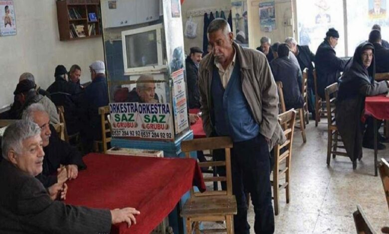 Busy cafe with red-tablecloths; elderly patrons chat while a man stands beside a pillar with an orchestra poster.